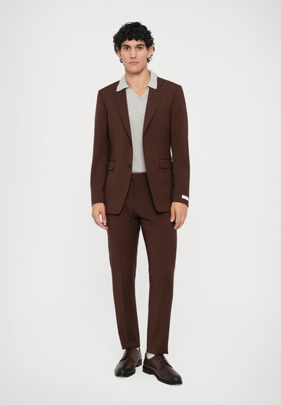 Costum maro croit cu pantaloni asortați, pullover gri cu decolteu în V dedesubt și pantofi eleganți închiși la culoare. Țesătură fină, design elegant și guler în contrast.