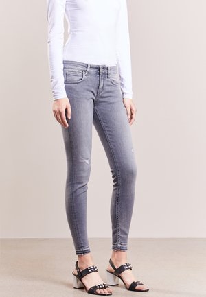 Vrouw draagt vervaagde grijze skinny jeans met lichte slijtage, een wit shirt met lange mouwen en zwarte sandalen met studs en blokhakken, staand.