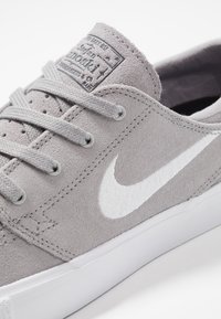 Graue Wildledersneaker mit weißem gesticktem Markenlogo, flachen Schnürsenkeln und einer weißen Gummisohle. Nahaufnahme betont Struktur und Design.