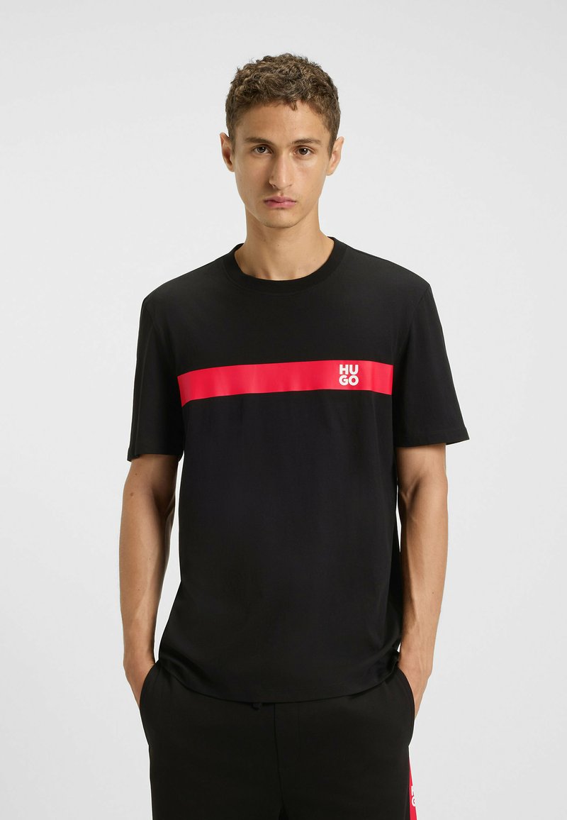 T-shirt nera in cotone con una striscia orizzontale rossa che presenta il logo "HUGO". Maniche corte e scollatura a girocollo.