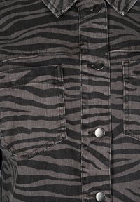 Grå denimjakke med et sort zebra-stribemønster, to brystlommer og sølvfarvede trykknapper. Teksturen ser robust og holdbar ud.