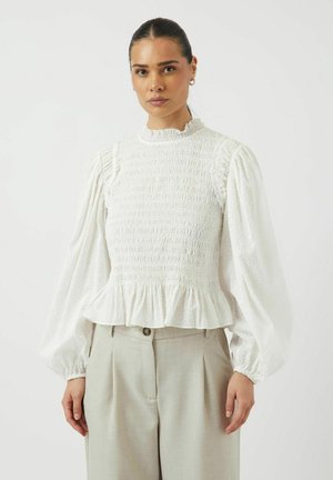 Blouse blanche avec une texture froncée et des manches longues bouffantes, dotée d'un ourlet volanté. Associée à un pantalon tailleur gris clair.