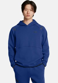Sudadera azul marino con un bolsillo tipo canguro, acentos negros en los hombros, un escote amplio y un logo sutil en el pecho. Textura de tela suave.