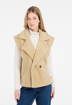 Gilet beige doppiopetto realizzato in tessuto morbido e testurizzato, con grandi revers e dettaglio dei bottoni, indossato sopra un top bianco a collo alto.