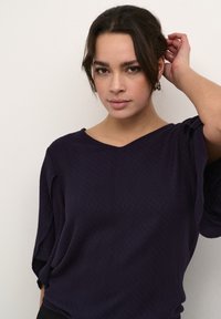 Lilla V-hals top med løse ærmer, som har et tekstureret diamantmønster. Stoffet ser blødt og let ud, med en afslappet pasform.