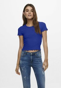 Blå kortad ribbad t-shirt med vågiga kanter, rund halsringning och korta ärmar. Kombinerad med slitna denimjeans.