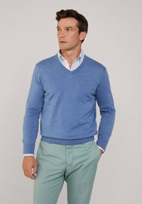 Maglione blu a scollo a V realizzato in un tessuto morbido, abbinato a una camicia a righe blu chiaro e pantaloni verde menta, con polsini e orlo a coste.