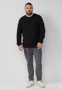 Zwarte gebreide trui met geribbelde textuur, ronde halslijn en lange mouwen. Gecombineerd met grijze cargobroek en witte sneakers.