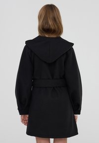 Donna con capelli castano chiaro di lunghezza alle spalle che indossa un cappotto nero con cappuccio e cintura, vista da dietro su uno sfondo semplice.