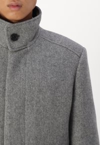 Cappotto in lana grigia con colletto alto, un bottone nero singolo e spalle strutturate, con una texture liscia e una vestibilità su misura.