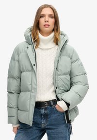 Lys grøn pufferjakke med hætte, med et quiltet design og sorte detaljer. Båret over en krämfarvet kabelstrikket sweater og blå jeans.