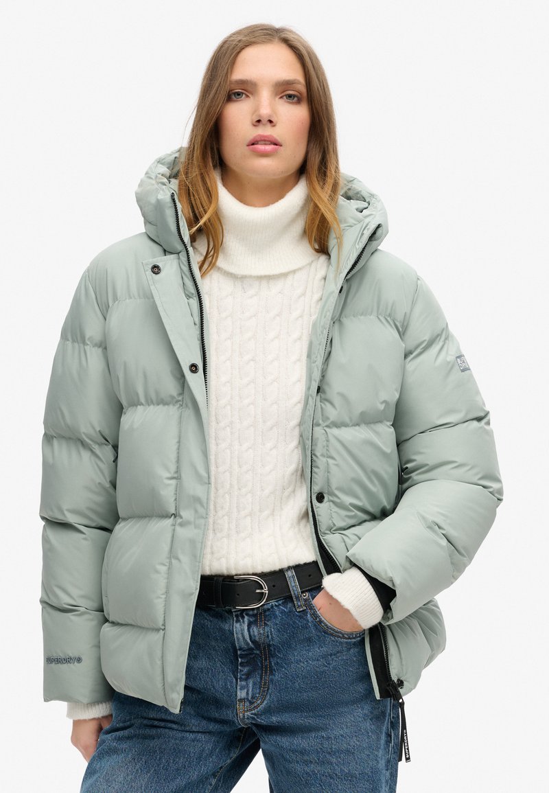 Lys grøn pufferjakke med hætte, med et quiltet design og sorte detaljer. Båret over en krämfarvet kabelstrikket sweater og blå jeans.