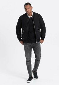 Svart puffjacka med dragkedjor, bärs över en svart polo, tillsammans med slitna grå jeans och svarta sneakers med vita sulor.