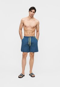 Mężczyzna bez koszulki, ubrany w niebieskie kąpielówki z żółtymi sznurkami i czarne klapki Moschino, stojący z rękami w kieszeniach na białym tle.