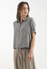 Camicia grigia a maniche corte con bottoni, tasca e orlo con cordino. Tessuto liscio con una leggera lucentezza.