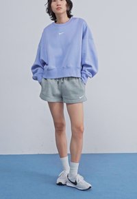 Oversized dukserica lavande boje s bijelim Nike logom, sivi šorti i bijele tenisice s crnim detaljima; glatka tkanina, opušteni kroj.