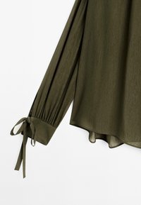 Blouse verte olive à manches longues avec poignets froncés noués en nœuds et un ourlet légèrement arrondi.