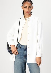 Cămașă din denim alb, cu nasturi în față, mâneci lungi, croială confortabilă, asortată cu blugi deschiși la culoare, cu talie înaltă și o geantă crossbody neagră.