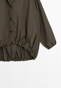 Veste vert olive foncé boutonnée avec poignets et ourlet élastiqués froncés, présentée sur un fond blanc.