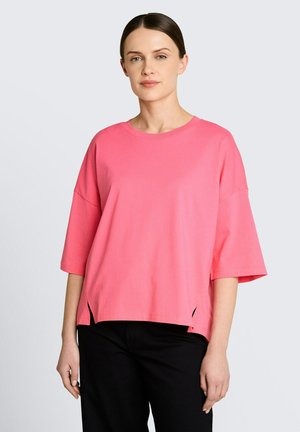 Vrouw draagt een losse roze korte mouwen shirt met zijsplitten, gecombineerd met een zwarte broek, staand tegen een effen lichte achtergrond.
