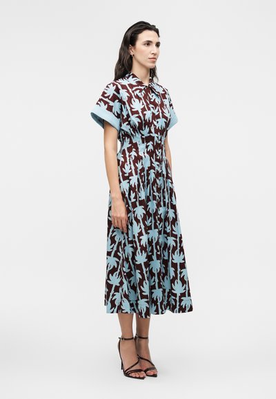 Γυναίκα που στέκεται, φορά φόρεμα μέχρι το γόνατο με μπλε λουλουδένια σχέδια σε σκούρο ύφασμα, κοντά μανίκια και μαύρα σανδάλια με ψηλό τακούνι.