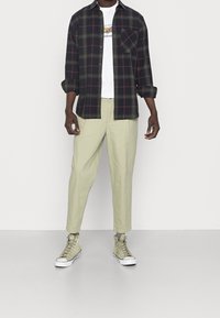 Man som bär beige byxor, ljusgröna hög-top sneakers, vit grafisk t-shirt och en oknäppt mörk rutig långärmad skjorta med uppvikta ärmar.