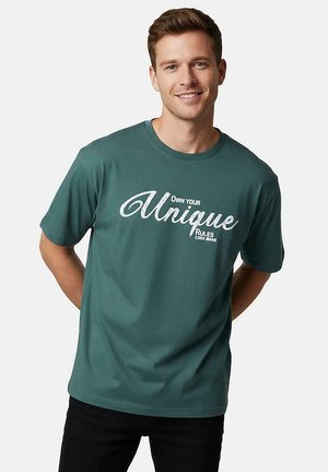 Lachende man die een groenblauw t-shirt draagt met de tekst "Own Your Unique Rules" op de voorkant, staand met handen achter de rug tegen een effen achtergrond.