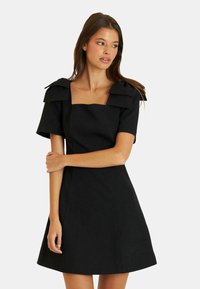 Schwarzes Kleid mit quadratischem Ausschnitt, kurzen Ärmeln und gefalteten Schulterakzenten. Hergestellt aus einem strukturierten Stoff mit tailliertem Oberteil und schwingendem Rock.