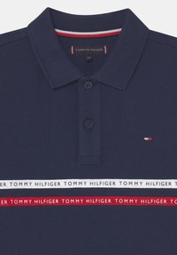 Tmavě modrá polo košile vyrobená z bavlny, s dvouknoflíkovým zapínáním, s texturovaným povrchem a logovými detaily v červených a bílých pruzích.