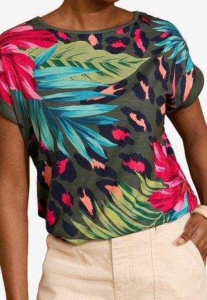 Chemise à manches courtes avec fond vert, imprimé floral tropical lumineux et imprimé animal foncé, portée avec un pantalon beige clair et un bracelet au poignet droit.