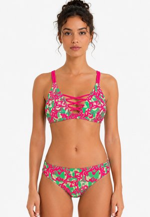 Woman wearing a pink and green abstract patterned bikini with crisscross straps on the top, standing against a white background.