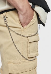 Hand in een beige cargobroekzak met een vastgemaakt zilveren ketting; persoon draagt een zwart-witte fleecedeken.