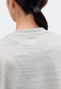 Personne aux cheveux foncés attachés en queue de cheval portant une chemise gris clair texturée, vue de dos sur un fond uni.