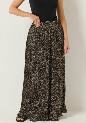 Mujer con una camiseta negra sin mangas y una falda larga plisada con estampado de leopardo, de pie con una mano en la cintura.