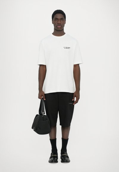 T-shirt en coton blanc avec du texte noir, associé à un short noir, des chaussettes noires et des mocassins noirs, tenant un petit sac noir.