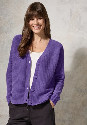 Lila gerippter Cardigan mit fünf Knöpfen, offenes Design und langen Ärmeln, getragen über einem weißen Top, vor einem neutralen Hintergrund.