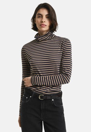 Woman with short dark hair wearing a beige and navy striped turtleneck and black jeans with a black belt, posing with one arm crossed.