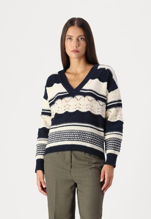 Maglione lavorato a maglia blu navy e crema con scollo a V, caratterizzato da varie righe e motivi testurizzati. Indossato con pantaloni su misura verde oliva.