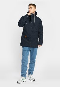 Navy blue insulated jacket with a hood, snap buttons, front pockets, and a brown label accent. Features a straight-cut design.