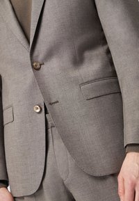 Jasnobrązowa marynarka wykonana z teksturowanej tkaniny, z dwoma guzikami i bocznymi kieszeniami. Marynarka ma dopasowany krój i czyste linie.