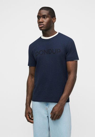 Άνδρας που στέκεται φορώντας μπλε ναυτικό T-shirt με την επιγραφή "DONDUP" πάνω από άσπρο εσώρουχο και ανοιχτόχρωμα τζιν, σε απλό φόντο.