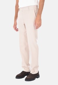 Pantaloni beige su misura con una texture liscia, caratterizzati da una chiusura con bottone e tasche laterali. Indossati con scarpe marroni scure.