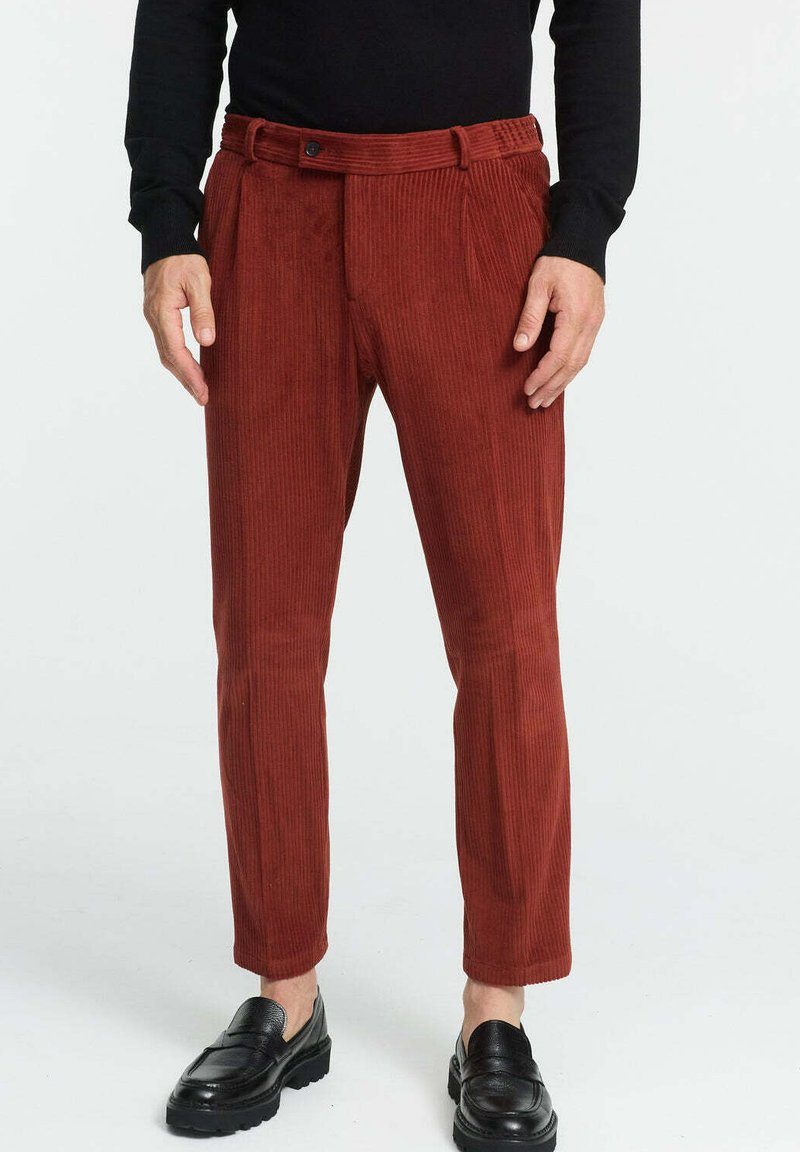 Kordhose in rostrot mit gerippter Textur, schmaler Schnitt und einem Bund mit Gürtelschlaufen. Stylisch kombiniert mit schwarzen Slippers.