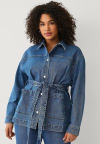 Jeanshemdjasje in medium blauw met een ceintuur, drukknoopsluiting en twee voorzakken. Contrasterende stiksels accentueren de naden.