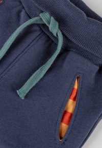 Die marineblauen Jogginghose verfügt über einen gestrickten Bund, eine geflochtene tealfarbene Kordel und karierte orange-rote Stoffe, die aus den Seitentaschen blitzen.