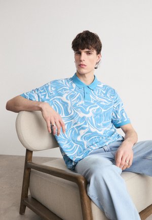 Sininen lyhythihainen polo, jossa on valkoinen pyörivä kuvio, ja siinä on kaulus sekä kolme nappia. Pukeutuneena vaaleansinisiin farkkuihin, istuu beigeillä tuoleilla.