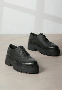 Chaussures Oxford en cuir noir avec une finition texturée, perforations décoratives et semelles en caoutchouc épaisses. Présentent un design classique à lacets.
