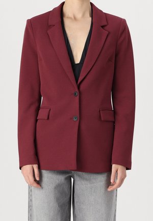 Personne portant un blazer bordeaux boutonné sur un haut noir et un jean gris clair, sur un fond clair uni.