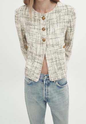 Kvinde iført en creme- og sort ternet cropped jakke med guldknapper og lyseblå højtaljede jeans mod en hvid baggrund.