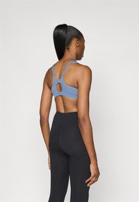 Gaiši zila sporta krūšturis ar plānām regulējamām siksnām un atslēgas caurumu detaļu aizmugurē, saskaņots ar augstiem melniem legingiem. Mīksta auduma tekstūra.